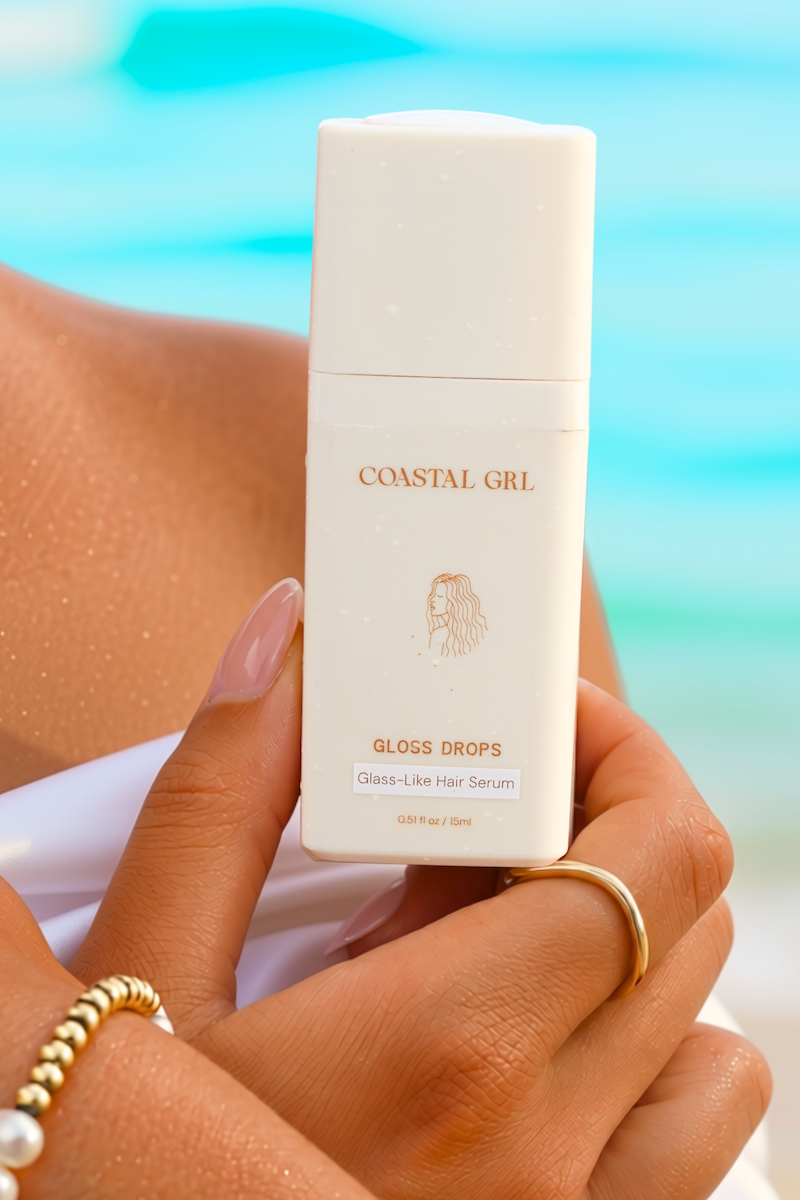 Hand holding a 15ml bottle of Coastal GRL Gloss Drops glass-like hair serum, with water droplets on the skin and a turquoise ocean behind.