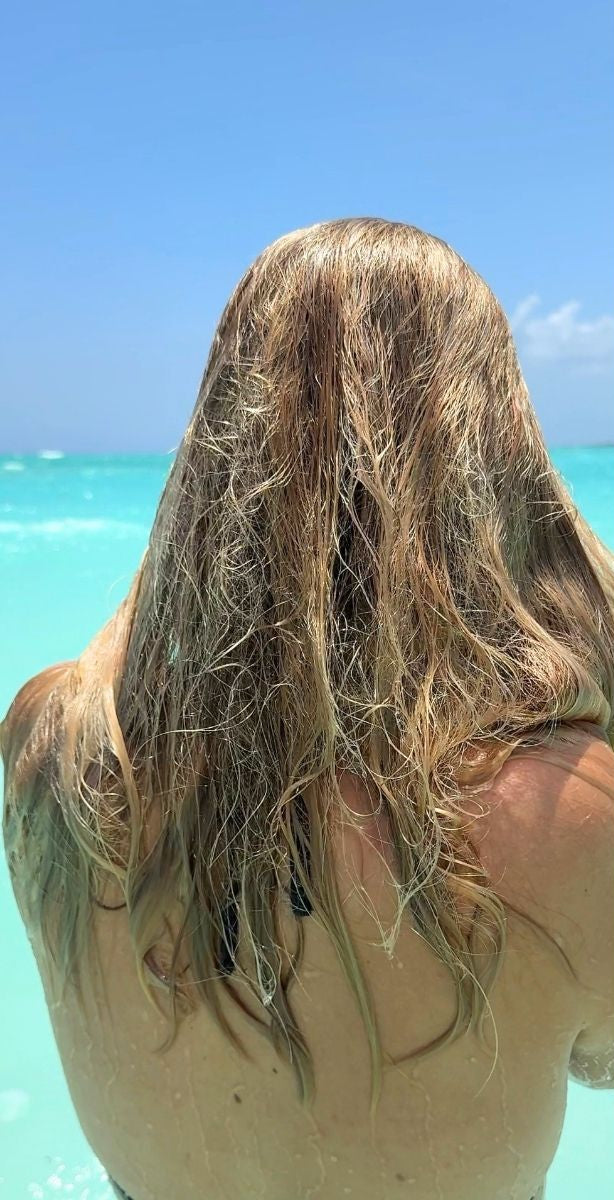 Before-and-after split screen showing a woman from behind at a beach, demonstrating the effect of a hair product. On the left, her hair is wet and frizzy.