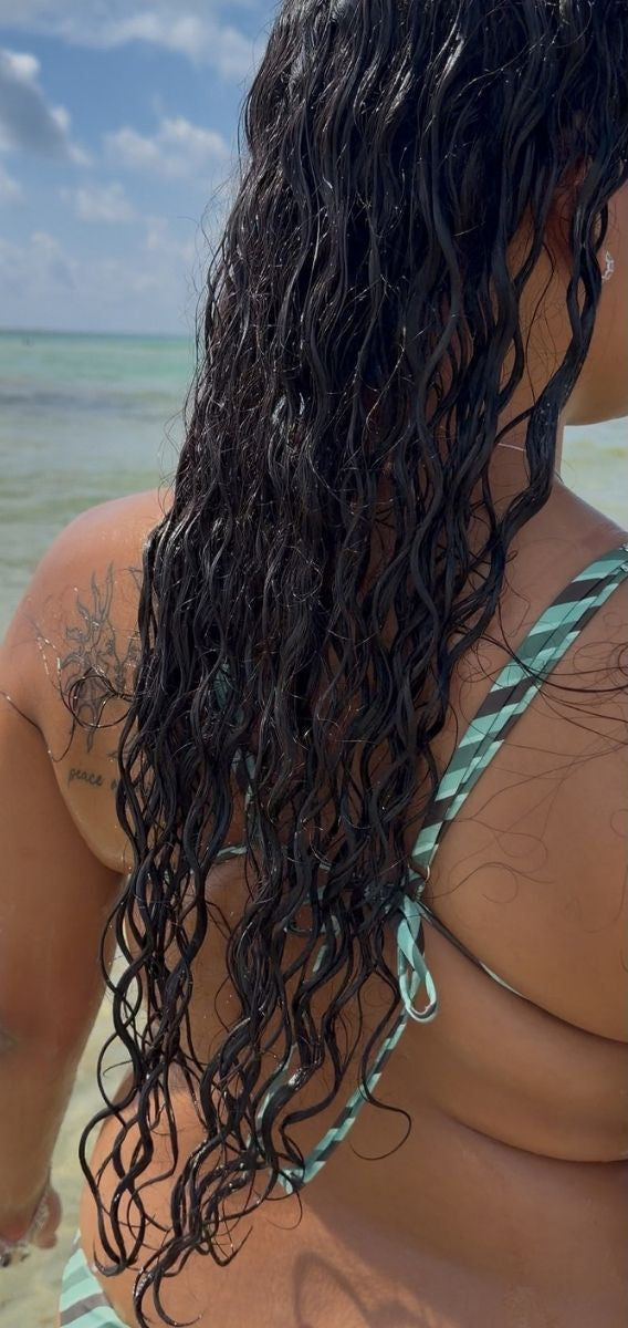 Side-by-side beach photo showing a woman from behind with very long, dark, curly hair in a green-and-white striped bikini. The left panel shows the hair wet, and the right panel shows it dry.