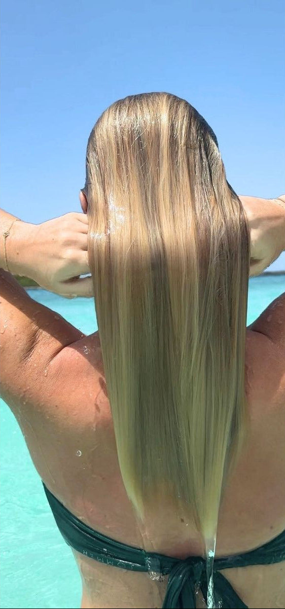 Before-and-after split screen showing a woman from behind at a beach, demonstrating the effect of a hair product. On the left, her hair is wet and frizzy.