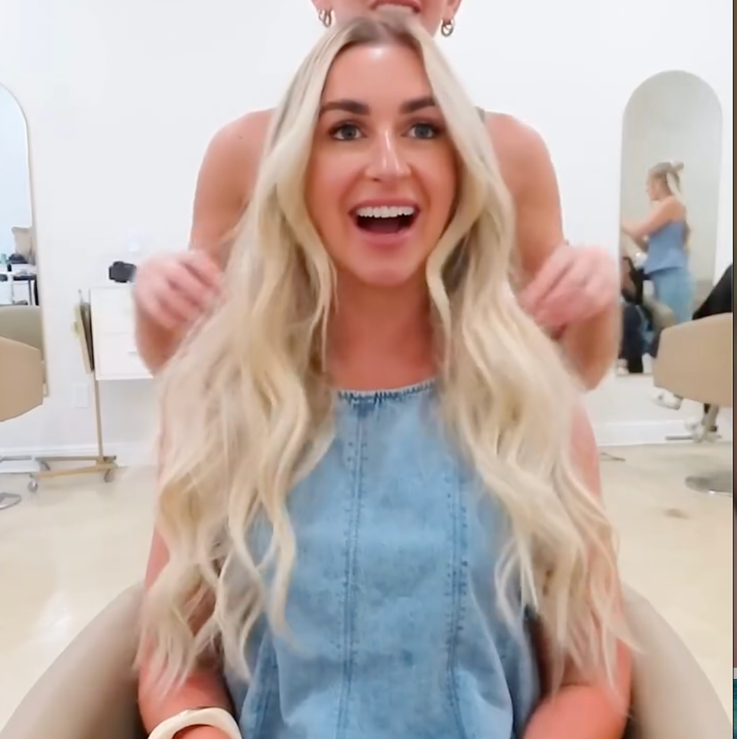 A woman with very long, wavy platinum blonde hair sits in a salon chair while another person stands behind her, holding and showing the hair. The setting is a bright, modern hair salon with mirrors and salon chairs visible in the background.