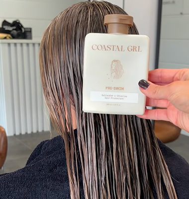 A person with long, wet brown hair sits with their back to the camera while a hand holds up a white bottle labeled 'COASTAL GRL PRE-SWIM' in front of their face. The setting appears to be a salon or beauty space with a white radiator and furniture in the background.